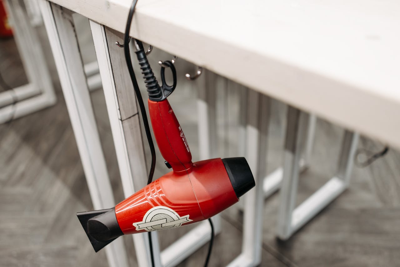 services-04 Close-up of a red hair dryer hanging from a white table indoors, showcasing product details.